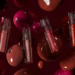 Transparent tubes of lip gloss float through the air surrounded by large, glossy, liquid-like droplets in rich shades of berry and mahogany. - Discover how Welpix and Art Director Zainab Sharrofna used CGI for cosmetics to create luxury visuals for Nice One, before products existed.