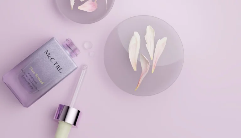 An open purple skincare bottle with its glass dropper resting on a lavender surface next to round glass dishes filled with white flower petals