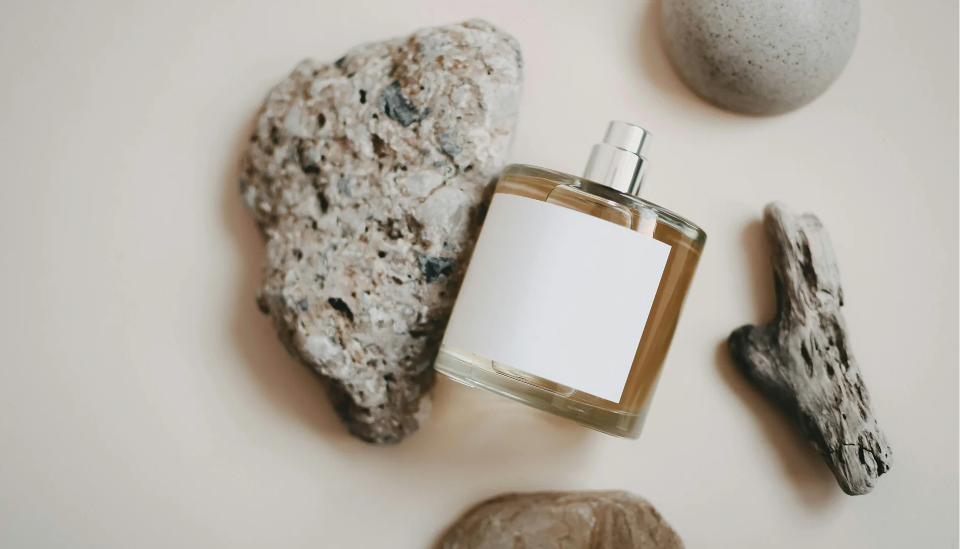 Top-down flat lay of a perfume bottle with a blank label, styled with natural props like rocks and driftwood on a neutral beige background.